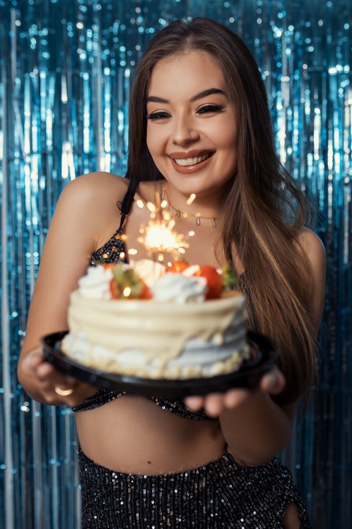 A happy woman holding a birthday cake with a lit sparkler against a festive background.